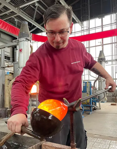 Glasmachr Ive Meusel steht nach vorne gebeugt und dreht mit der linken Hand die Glasmacherpfeife mit dem heißen Glasposten. Die rechte Hand hält einen großen hölzernen Löffel mit dem er das rot-glühenden Glas formt. Ive trägt einen roten für die ELIAS Glashütte typischen Sweatert und steht neben dem Wasserbasin zum Küheln des Löffels.