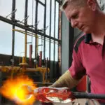 Glasmacher Ricardo Scholz ist von vorne zu sehen. Er trägt einen rotes für die ELIAS Glashütte typisches Polo und hält mit der linken Hand die Glasmacherpfeife an der eine Glasmurmel vorgeformt ist. Mit der rechten Hand wärmt er mit einem Handbrenner die Murmel mit großer und scharfer Flamme durch. Im Hitergrund sind die große Glasfenster des Hüttenbereichs der ELIAS Glashütte zu sehen.