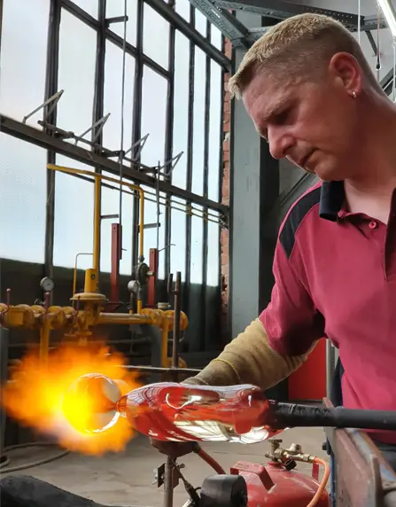 Glasmacher Ricardo Scholz ist von vorne zu sehen. Er trägt einen rotes für die ELIAS Glashütte typisches Polo und hält mit der linken Hand die Glasmacherpfeife an der eine Glasmurmel vorgeformt ist. Mit der rechten Hand wärmt er mit einem Handbrenner die Murmel mit großer und scharfer Flamme durch. Im Hitergrund sind die große Glasfenster des Hüttenbereichs der ELIAS Glashütte zu sehen.