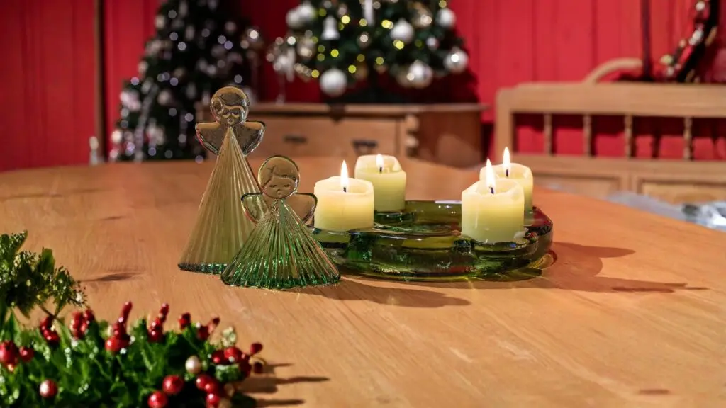 Je 1 kleiner und großer grüner Glasengel und ebenso ein grüner Adventskranz aus Thüringer Waldglas mit brennenden Stumpenkerzen auf einem hellen Holztisch. Im Hintergrund geschmückte, beleuchtete Weihnachtsbäume sowie eine rote Holzwand. Der Hintergrund erscheint komplett mit Bokeh.
