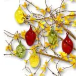 Strauss mit gelben Blüten und farbigen Glas-Ostereiern (grün, gelb, rot) vor weißem Hintergrund.